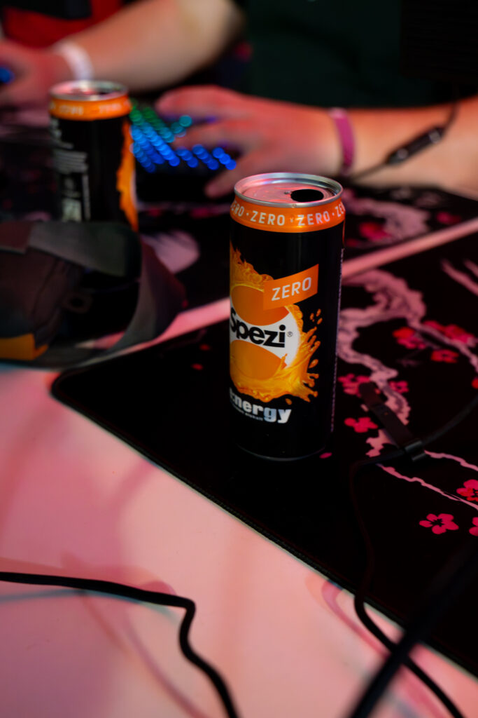 Eine Dose Spezi Energy Zero steht auf einem Gaming-Schreibtisch neben Tastatur, Mauspad und Headset. Im Hintergrund sind unscharf die Hände eines Spielers an Maus und Tastatur zu erkennen.