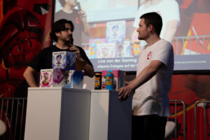 Zwei Moderatoren stehen auf der Gaming Stage der DoKomi und führen ein Gespräch. Auf den Podesten vor ihnen stehen League-of-Legends-Figuren, Spezi-Dosen und weitere Gewinne, während im Hintergrund die Live-Übertragung auf einer großen Leinwand zu sehen ist.
