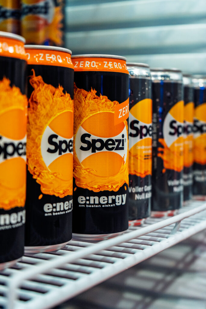 Mehrere gekühlte Dosen Spezi Energy Zero stehen in einer Reihe auf einem Kühlschrankregal. Die orange-schwarzen Dosen sind im Vordergrund scharf abgebildet, während weitere Getränke unscharf im Hintergrund zu sehen sind.