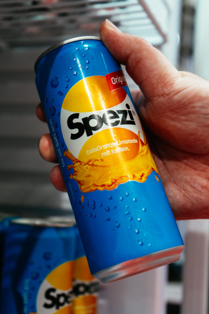 Eine Hand hält eine gekühlte Dose Spezi Original vor einem Getränkekühlschrank. Die Cola-Orange-Limonade ist im Fokus, während weitere Dosen im Hintergrund verschwimmen.
