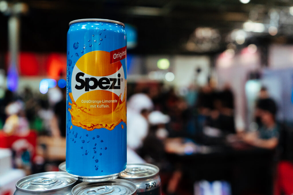 Eine einzelne Dose Spezi Original steht erhöht auf mehreren Dosen vor einem unscharfen Messehintergrund. Die Aufnahme hebt den neuen Vereinspartner von eSports Cologne in den Mittelpunkt.
